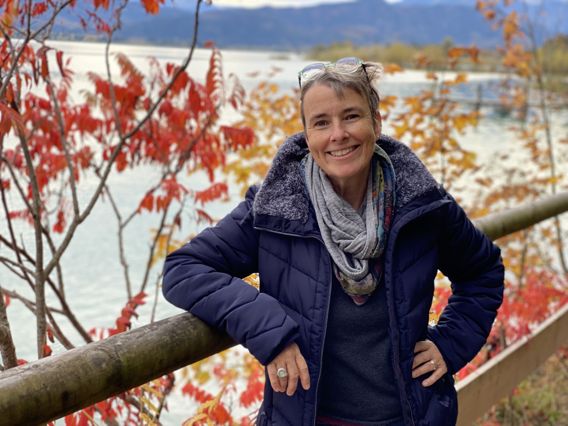 Das Foto zeigt Carola Lehmann. Sie lehnt sich dabei an ein Geländer an. Im Hintergrund ist ein See zu sehen sowie Bäume mit buntem Herbstlaub.