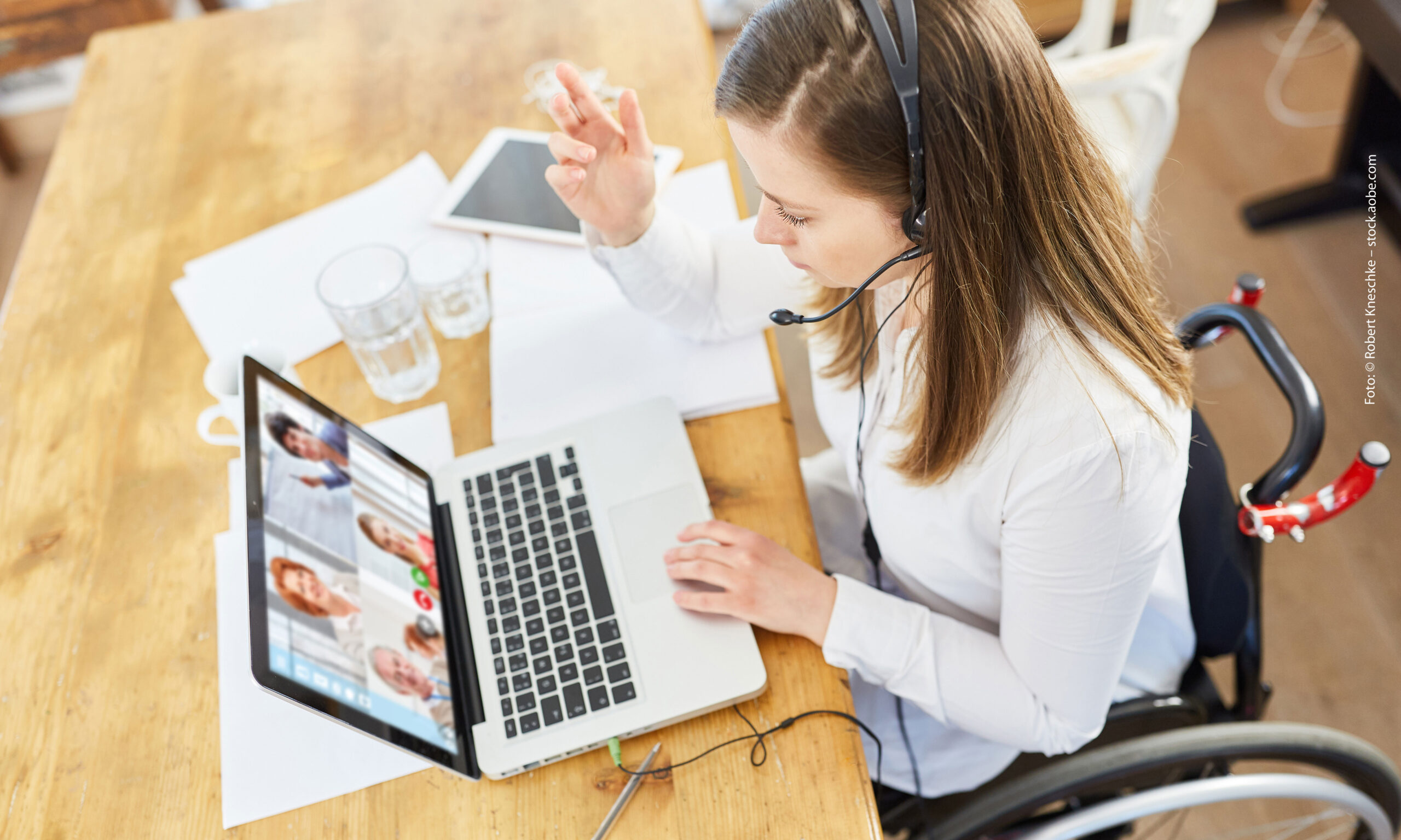 Eine Frau im Rollstuhl sitzt an einem Tisch und nimmt an einem Online-Meeting teil. Foto: © Robert Kneschke – stock.adobe.com